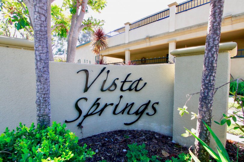 A WHITE COLOUR WALL THAT REPRESENTS VISTA SPRINGS AND THE BUILDING BEHIND THE WALL SOME TREES ALSO AVAILABLE IN FRONT OF THE WALL.